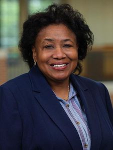 A woman with curly black hair smiles while wearing a blue blazer over a striped shirt against a blurred background.