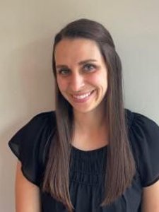 A woman with long straight brown hair wearing a black blouse smiles while standing against a plain light-colored wall.
