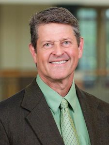 A middle-aged man with short brown hair smiles wearing a green shirt, patterned tie, and dark brown blazer indoors.