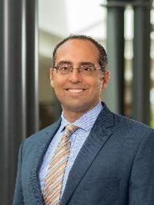 A man wearing glasses, a blue suit, striped tie, and checkered shirt stands smiling in a corridor with black columns.