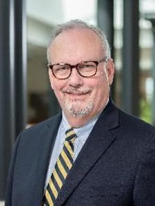 A middle-aged man with glasses and a short gray beard smiles while wearing a dark suit and striped tie indoors.