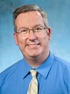A middle-aged man with short gray hair and glasses smiles while wearing a blue shirt and yellow striped tie against a blue.