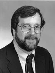 A black and white portrait of a man with glasses, a beard, and a suit jacket smiling slightly at the camera.