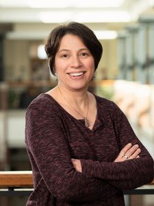 A person with short dark hair smiles with arms crossed wearing a maroon long-sleeve shirt and necklace indoors.