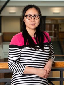 A woman with long dark hair and glasses stands indoors leaning on a wooden railing, wearing a striped shirt with a pink top.