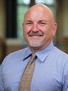A bald man with a goatee and hoop earring smiles wearing a blue shirt and patterned tie in a blurred indoor setting.