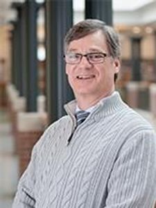 A man wearing glasses and a light gray cable-knit sweater smiles while standing indoors with blurred architectural elements.