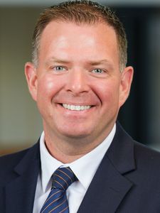 A man with short brown hair wearing a dark suit, white shirt, and striped tie smiles at the camera.