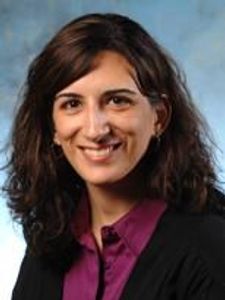 A woman with shoulder-length dark brown hair smiles at the camera wearing a purple blouse and black cardigan.