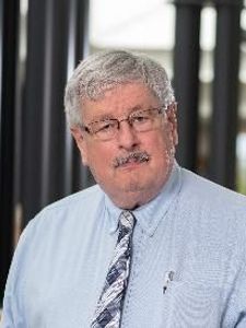 A man with gray hair and glasses wearing a light blue shirt and patterned tie stands in front of black columns outdoors.