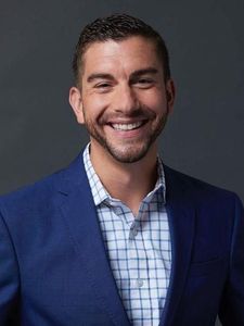 A man with short dark hair and a beard smiles while wearing a blue suit jacket and a white checkered shirt.