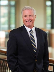 A smiling older man with gray hair wearing a dark suit and striped tie stands indoors near a railing.