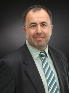 A man wearing a dark checkered suit jacket, white shirt, and black lanyard stands against a dark gray background.