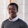 JCU faculty member, a man with dark skin and a warm smile, poses for his portrait in a grey sweater and white shirt.