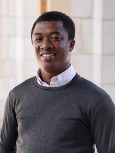 JCU faculty member, a man with dark skin and a warm smile, poses for his portrait in a grey sweater and white shirt.