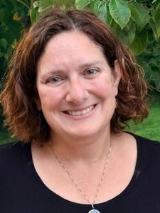 A JCU faculty member with curly brown hair and a bright smile wears a black top and a silver necklace