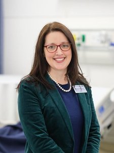 A woman with long brown hair and red glasses smiles while standing in a room with a hospital bed in the background.