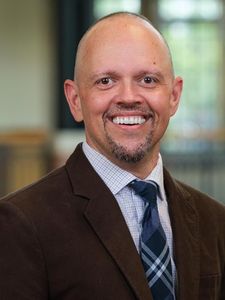 A smiling bald man with a goatee wearing a brown jacket, checkered shirt, and blue plaid tie stands indoors.