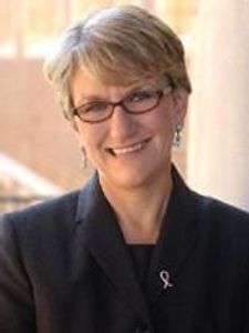 A smiling woman with short blonde hair and glasses wears a dark blazer and a pink ribbon pin while standing indoors.
