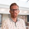 A man with short red hair, glasses, and a beard stands indoors wearing a white polo shirt with a small pattern.
