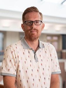 A man with short red hair, glasses, and a beard stands indoors wearing a white polo shirt with a small pattern.
