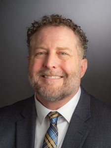 A middle-aged man with curly hair and a beard smiles wearing a dark suit, white shirt, and striped tie against a gray.