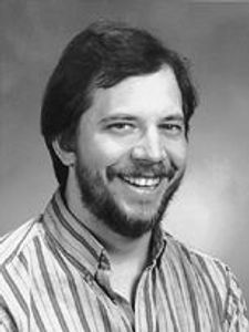 A man with dark hair and a beard smiles while wearing a striped button-up shirt against a plain gray background.