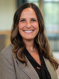 A woman with long wavy brown hair smiles at the camera wearing a black blouse and houndstooth blazer.