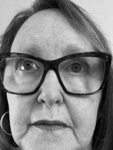 Close-up black and white photo of an older woman wearing large rectangular glasses and hoop earrings looking slightly upward.