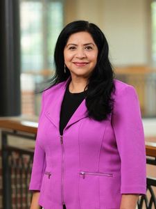 A woman with long black hair wearing a bright purple jacket smiles while standing indoors near a railing.