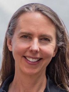 A woman with long light brown hair smiles gently against a plain blue-gray background.