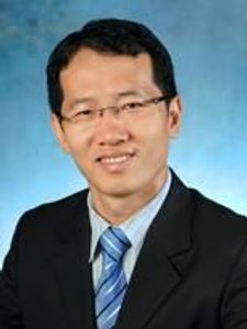 A man wearing glasses, a black suit, white shirt, and blue striped tie smiles against a blue studio background.