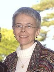 A person with short gray hair and glasses smiles outdoors wearing a white turtleneck and a multicolored jacket.