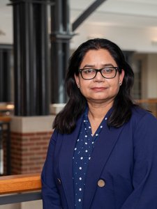 A JCU faculty member with dark hair and glasses, wearing a blue blazer, stands looking at the camera in a university atrium.