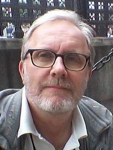 A middle-aged man with light brown hair, glasses, and a short beard wearing a checkered shirt against a blue background.
