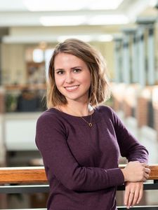 A woman with shoulder-length light brown hair smiles while leaning on a wooden railing in a bright indoor space.