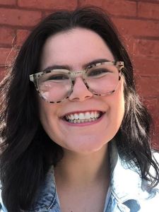 A person with dark hair and tortoiseshell glasses smiles broadly in front of a red brick wall while wearing a light denim.