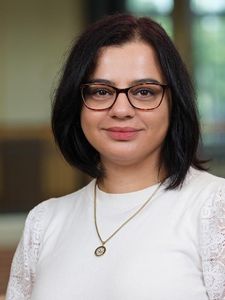 A woman with shoulder-length dark hair and tortoiseshell glasses smiles softly wearing a white top and a round pendant.