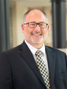 A middle-aged man with glasses and a trimmed beard smiles while wearing a dark suit and checkered tie indoors.