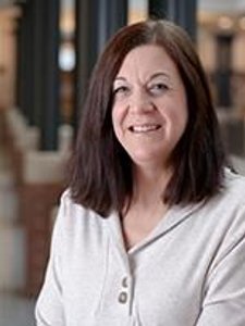 A woman with shoulder-length brown hair wearing a light gray buttoned shirt smiles while seated indoors.