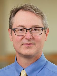 A man with short gray hair and glasses wears a blue shirt and yellow patterned tie against a blurred background.