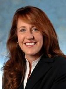 A woman with long reddish-brown hair wearing a black blazer and white shirt smiles against a blue-gray background.
