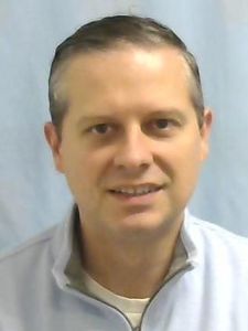 A man with short brown hair wearing a light gray quarter-zip sweater smiles slightly against a plain light blue background.