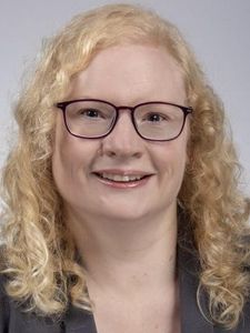 A person with curly blonde hair and glasses smiles while wearing a dark blazer against a plain background.