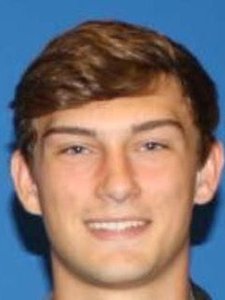 A young man with short brown hair smiles slightly against a solid blue background.