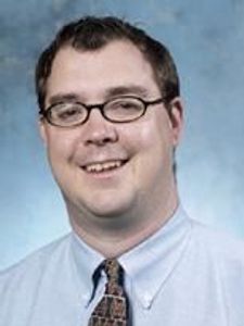A man with short dark hair and glasses smiles wearing a light blue shirt and patterned tie against a blue-gray background.