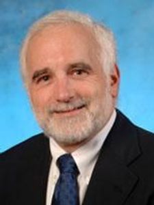 A smiling older man with white hair and a beard wearing a dark suit, white shirt, and blue tie against a blue background.
