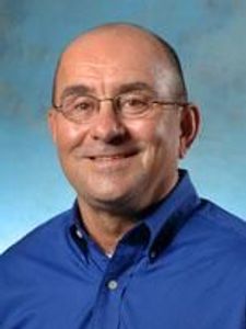 A smiling bald man wearing glasses and a blue button-up shirt poses against a blue and green gradient background.