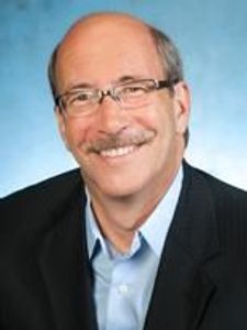 A middle-aged man with glasses and a mustache smiles while wearing a dark blazer and light blue shirt against a blue.