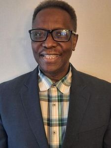 A man wearing black glasses, a dark blazer, and a plaid shirt smiles slightly against a plain wall.